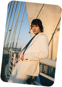 Man in a white suit leaning on a bridge railing with cables in the background at sunset.