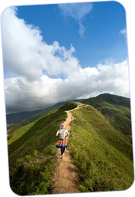 Hiker walking along a narrow dirt trail on a green mountain ridge under a blue sky with scattered clouds.