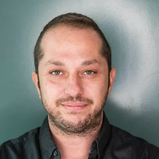 Headshot of a man with short hair and light facial hair wearing a dark shirt against a teal background.