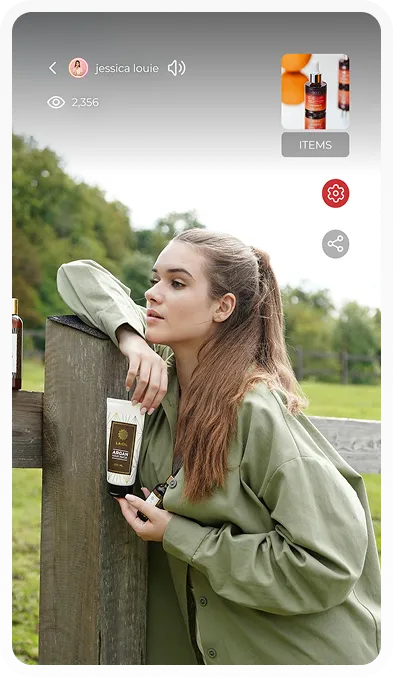 Woman with long brown hair leaning on a wooden fence holding a tube of argan hand cream outdoors.