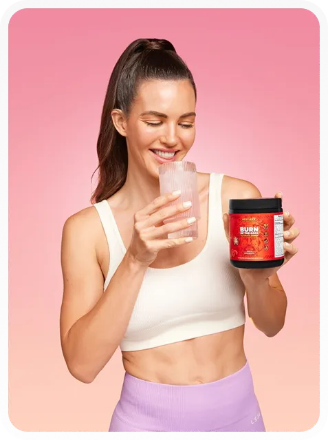 Smiling woman in white sports bra and purple leggings holding a glass of water and a red container of Burn supplement against a pink background.