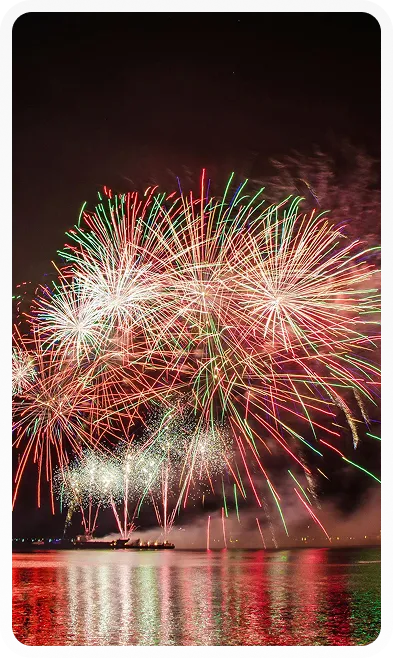 Colorful fireworks bursting over calm water at night with reflections on the surface.