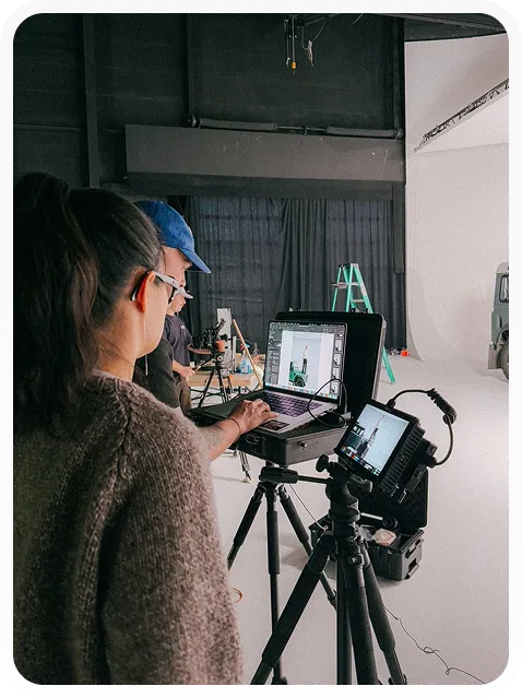 Two people working in a photo studio with camera equipment and a laptop showing product images.