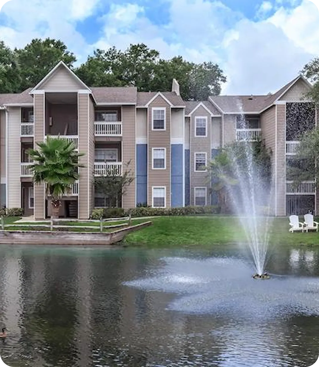 Apartment complex with three-story buildings facing a pond with a water fountain and green lawn.