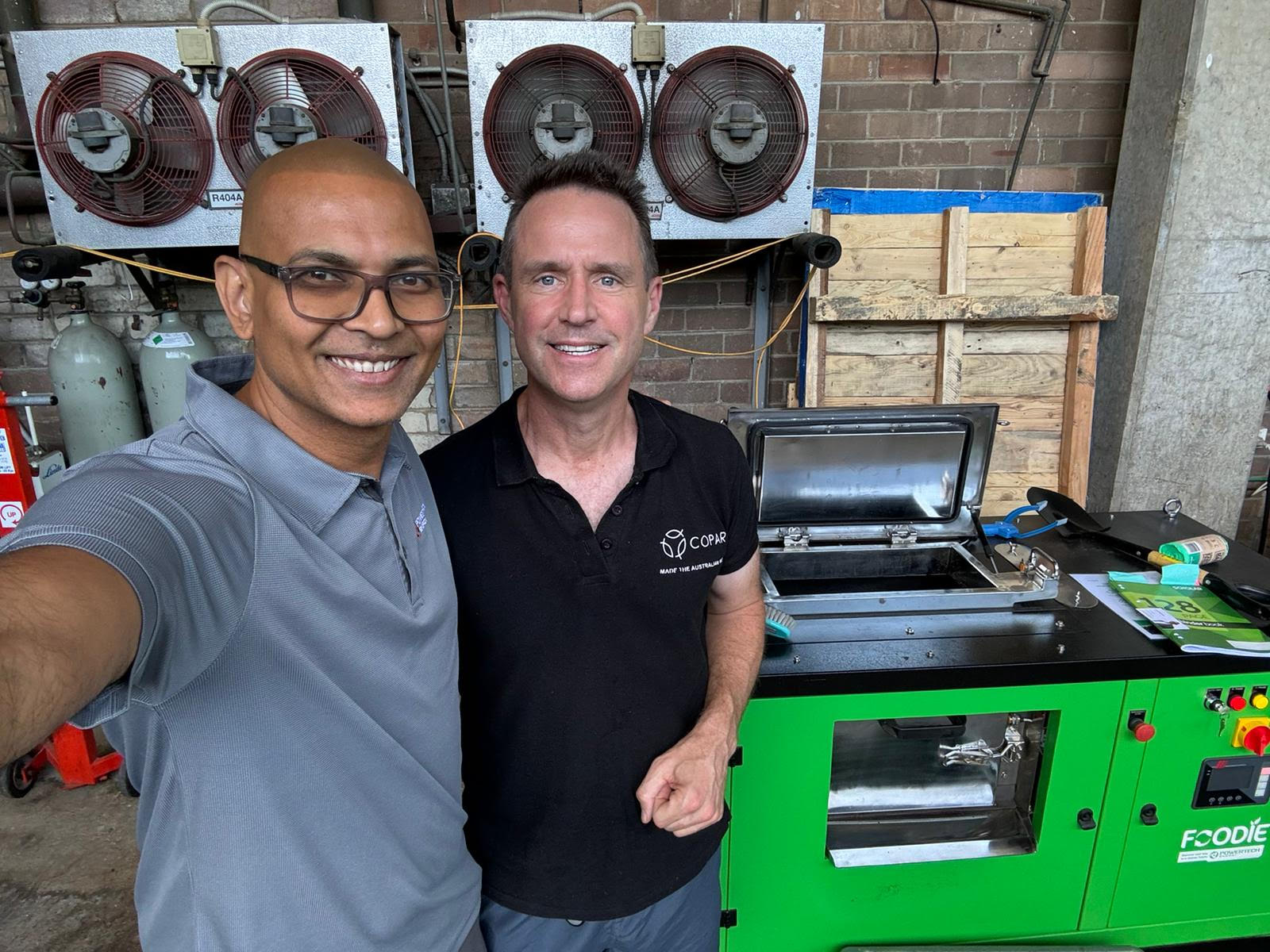 Two men standing next to a cmmercial food waste composter machine. The background shows industrial equipment, including organic waste composter and gas cylinders