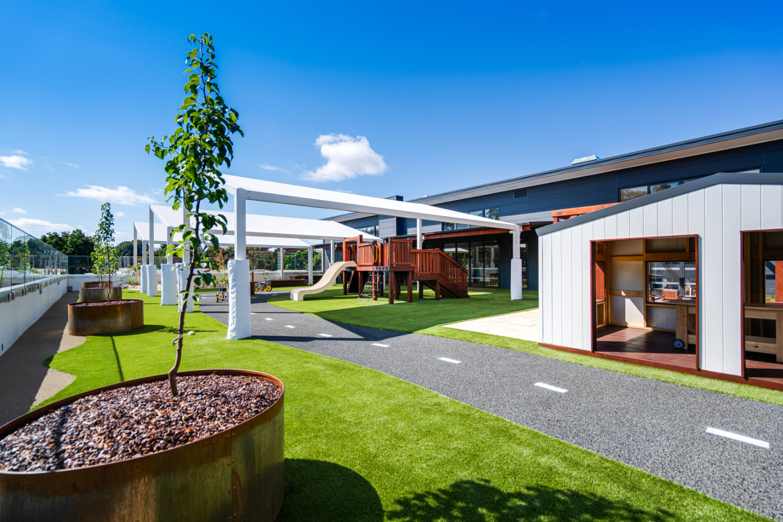 Outdoor playground with artificial grass, small trees in circular planters, wooden play structure with slide, and a playhouse under a clear blue sky.