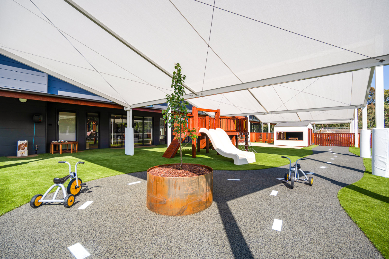 Outdoor children's playground with a white slide, wooden play structure, two tricycles on a marked pathway, and a small tree planted in a round metal planter under large shade sails.