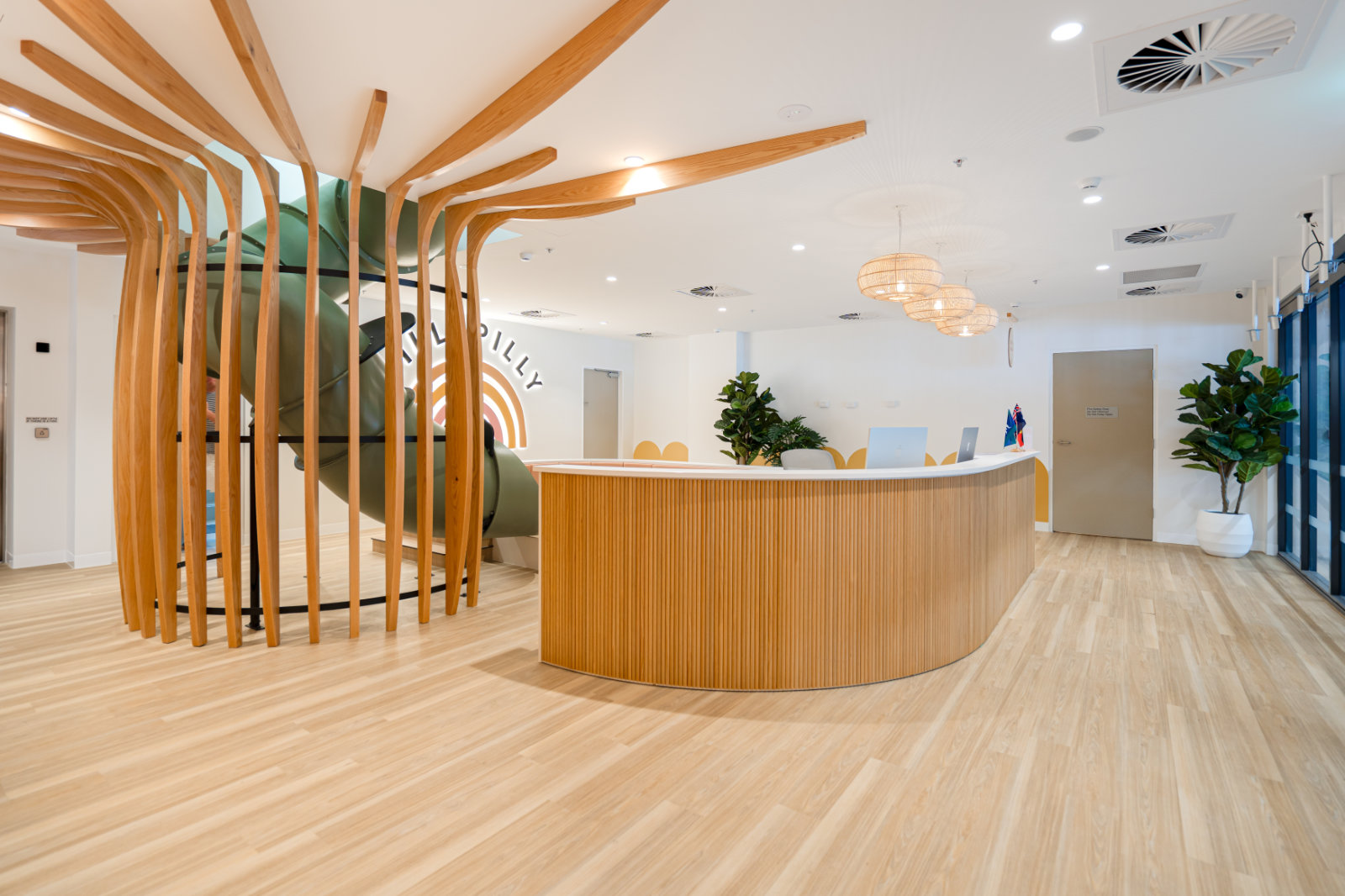 Modern wooden reception desk in a bright room with a green slide enclosed by vertical wooden slats and decorative plants.
