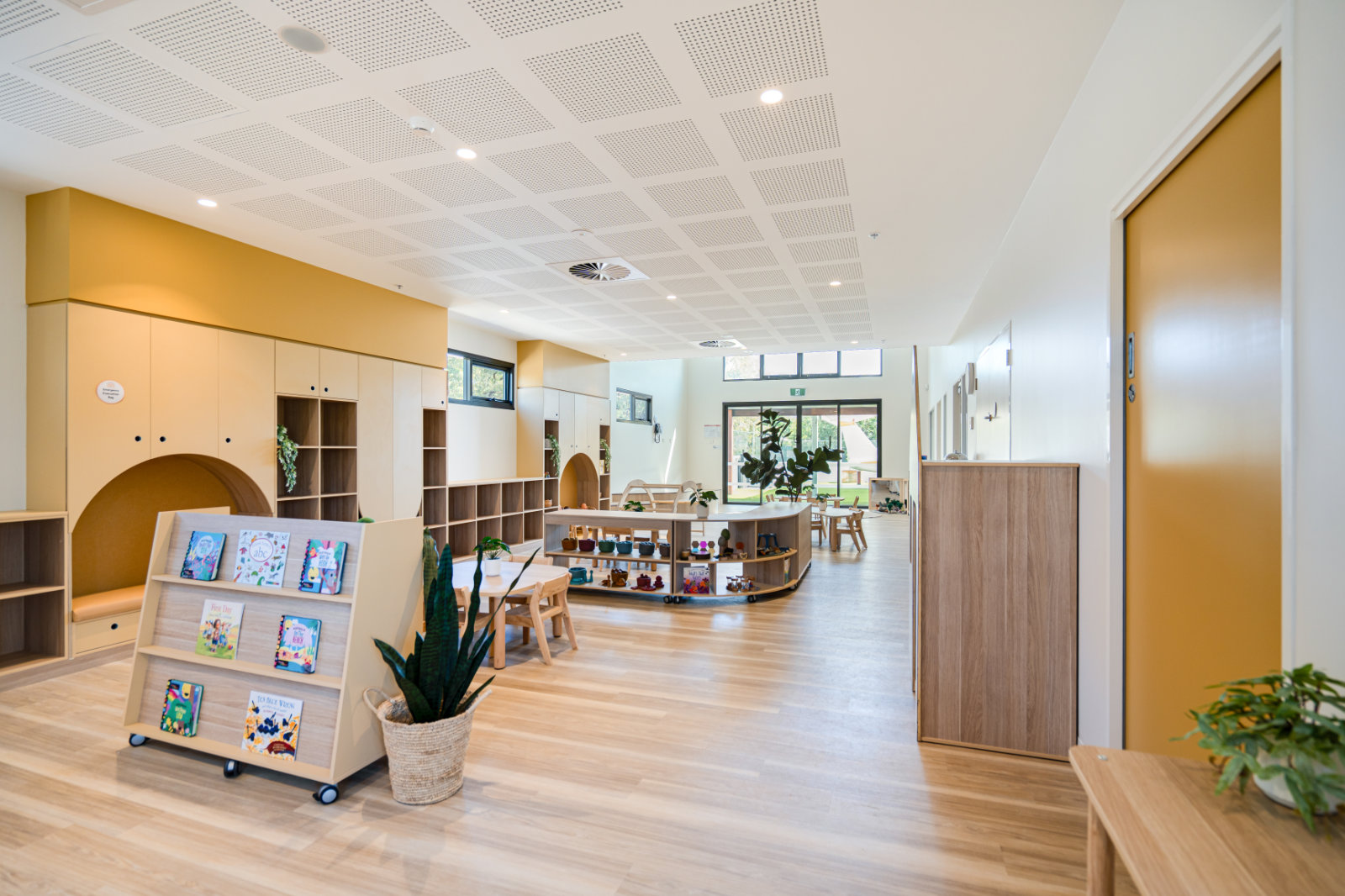 Bright, modern classroom with wooden floors, mustard yellow cabinets, low bookshelves, child-sized tables and chairs, and plants.