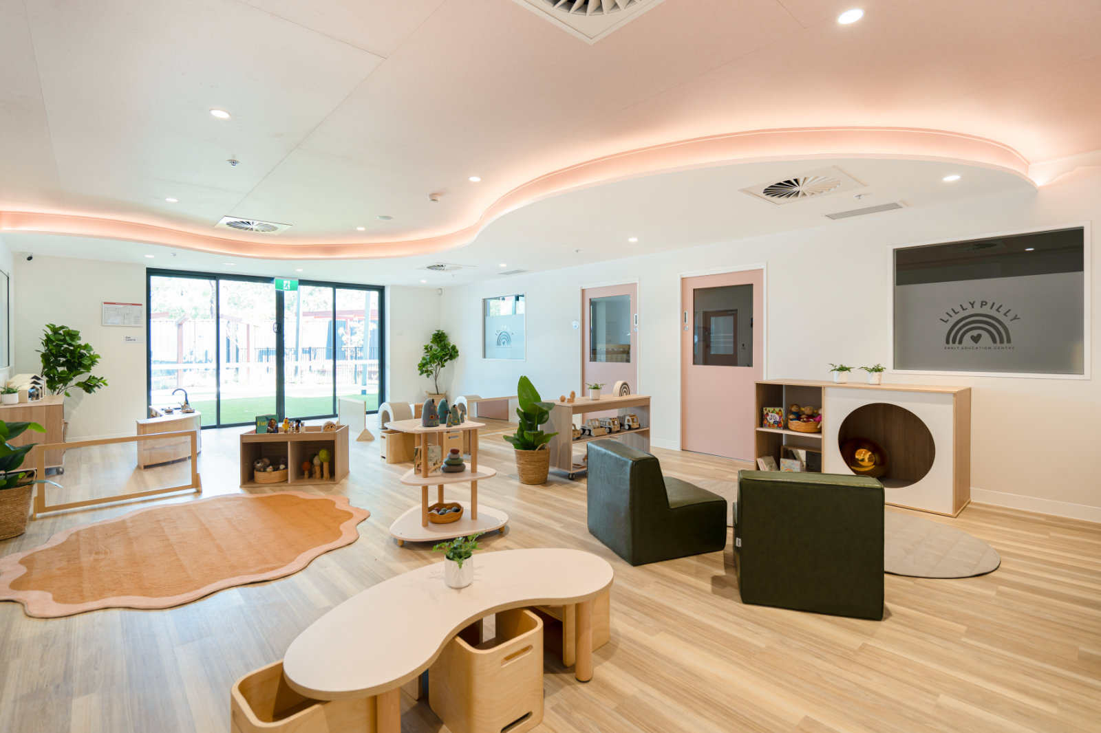 Bright, modern early education classroom with wooden furniture, green chairs, plants, toys, and large glass doors leading outside.