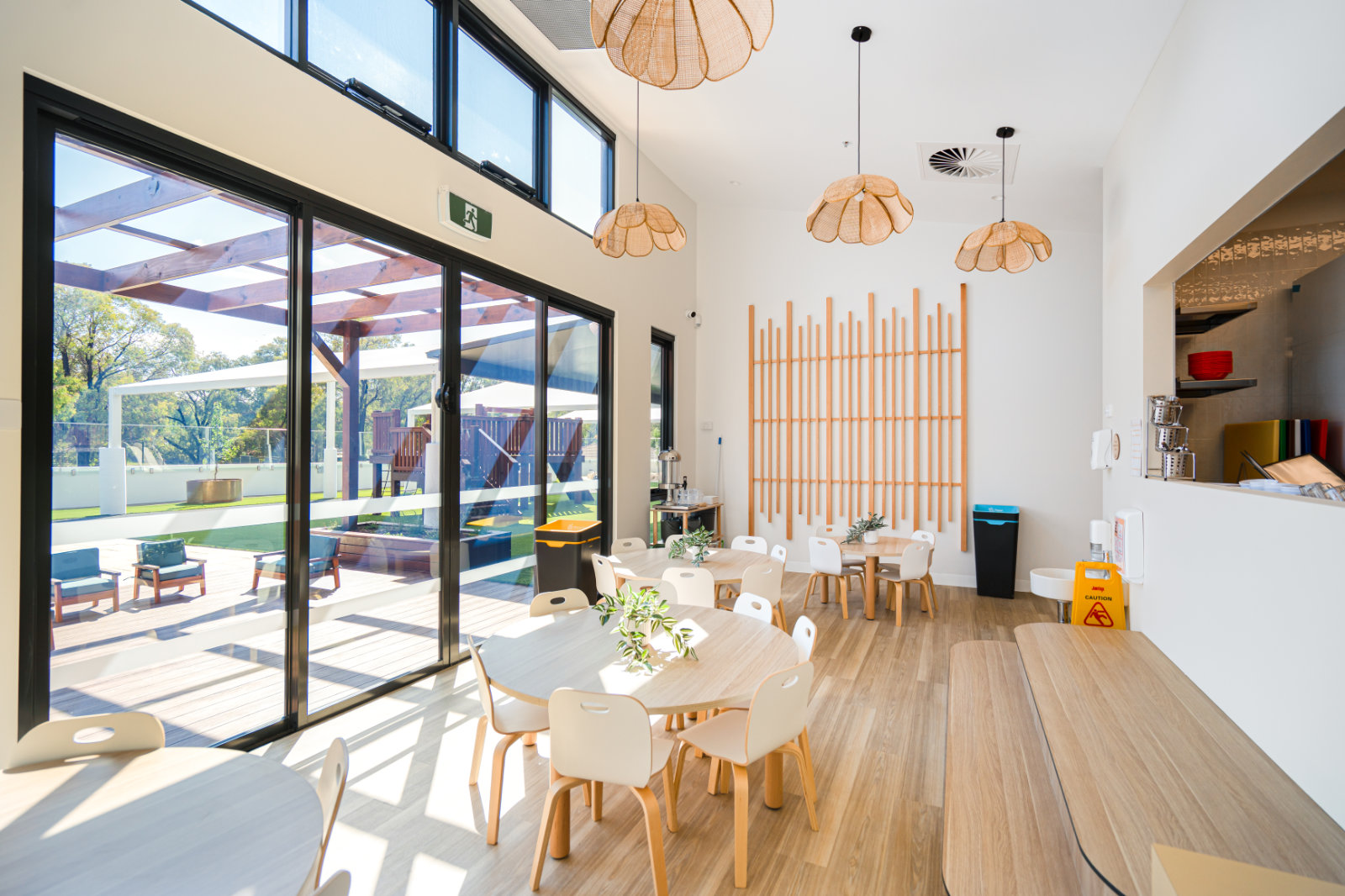 Bright modern dining area with round wooden tables and white chairs, hanging wicker lamps, and large glass doors opening to an outdoor patio with seating and greenery.