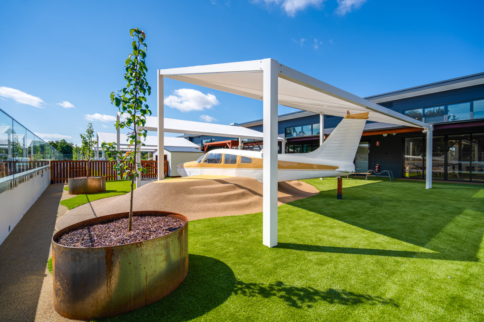 Small white and gold airplane mounted on a green artificial turf lawn under white shade structures near a modern building and metal planters with young trees.