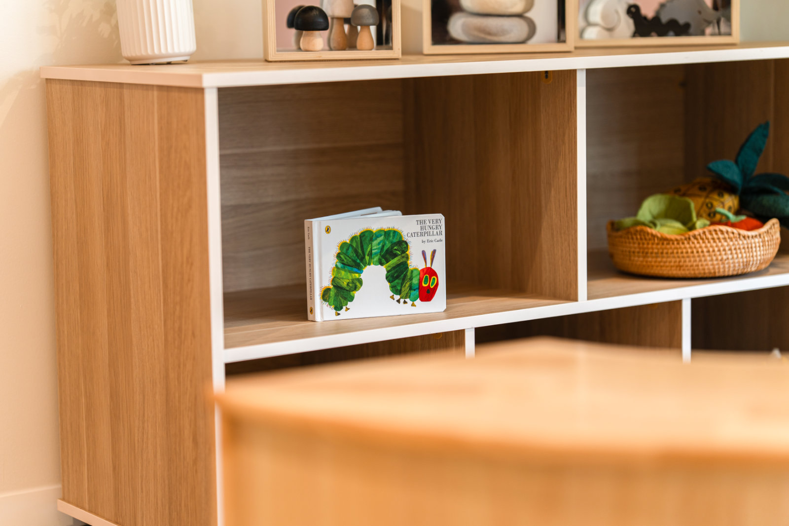 The children's book 'The Very Hungry Caterpillar' displayed on a wooden shelf with other toys and decorations.