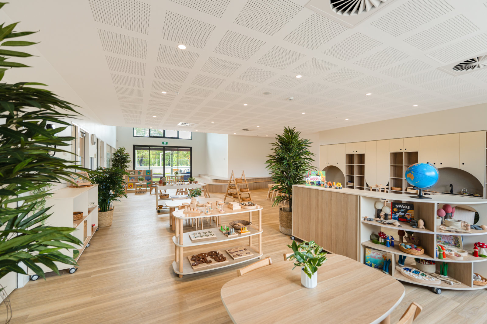 Bright, spacious early childhood classroom with wooden tables, shelves with educational toys, a globe, and indoor plants.