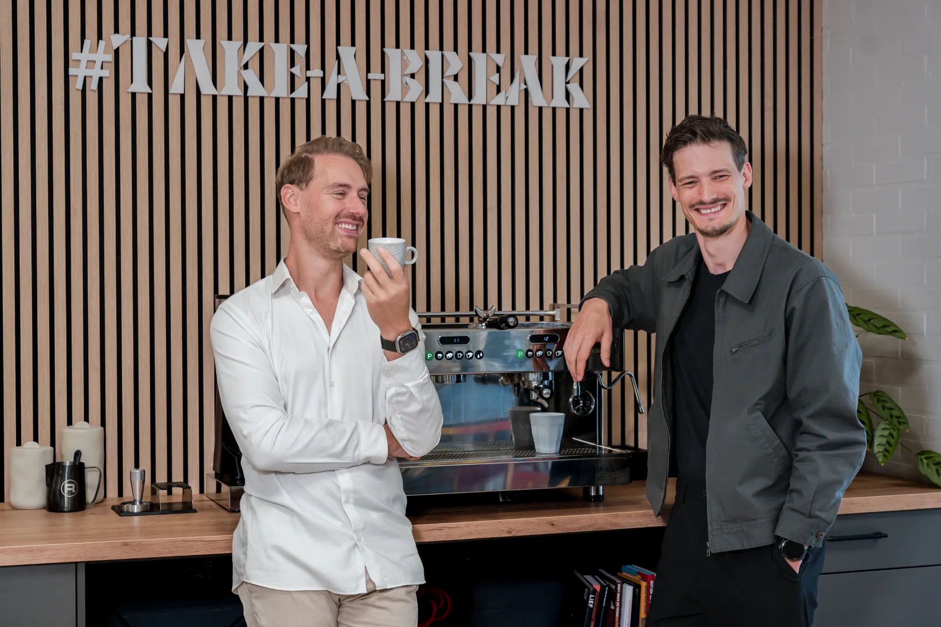 Twee collega's van De Zakerij lachend bij de koffiebar onder #TAKE-A-BREAK