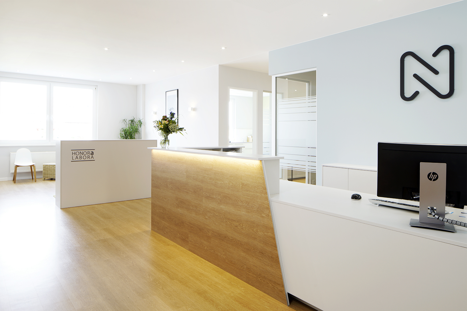 Moderner Empfangsbereich mit Holzboden, weißen Schreibtischen, einer Blumenvase, einem Computerbildschirm und viel Tageslicht durch große Fenster.