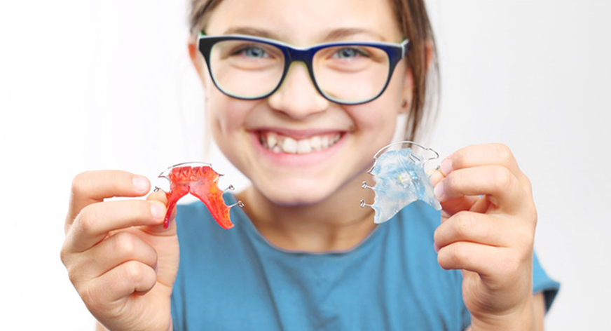 Lächelndes Mädchen mit blauer Brille und blauem Shirt, das zwei herausnehmbare Zahnspangen hält – eine rote und eine transparente.