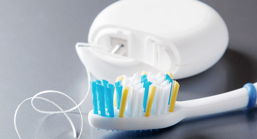 Nahaufnahme einer weißen Zahnbürste mit blauen und gelben Borsten neben einer Dose mit Zahnseide auf grauer Oberfläche.