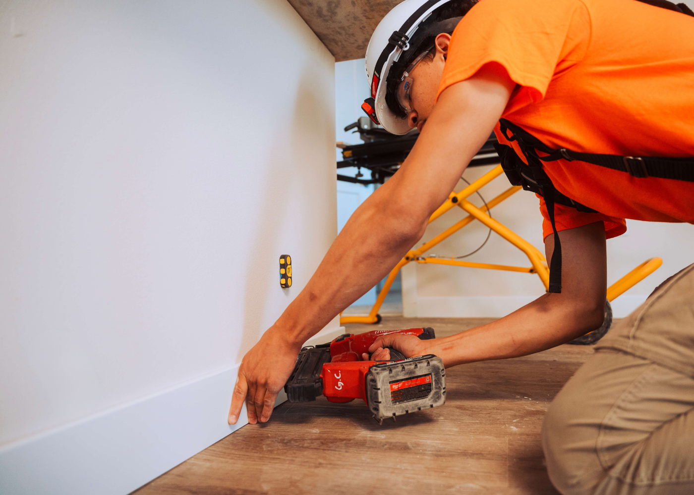 A carpenter uses a nail gun to apply wood baseboard. 