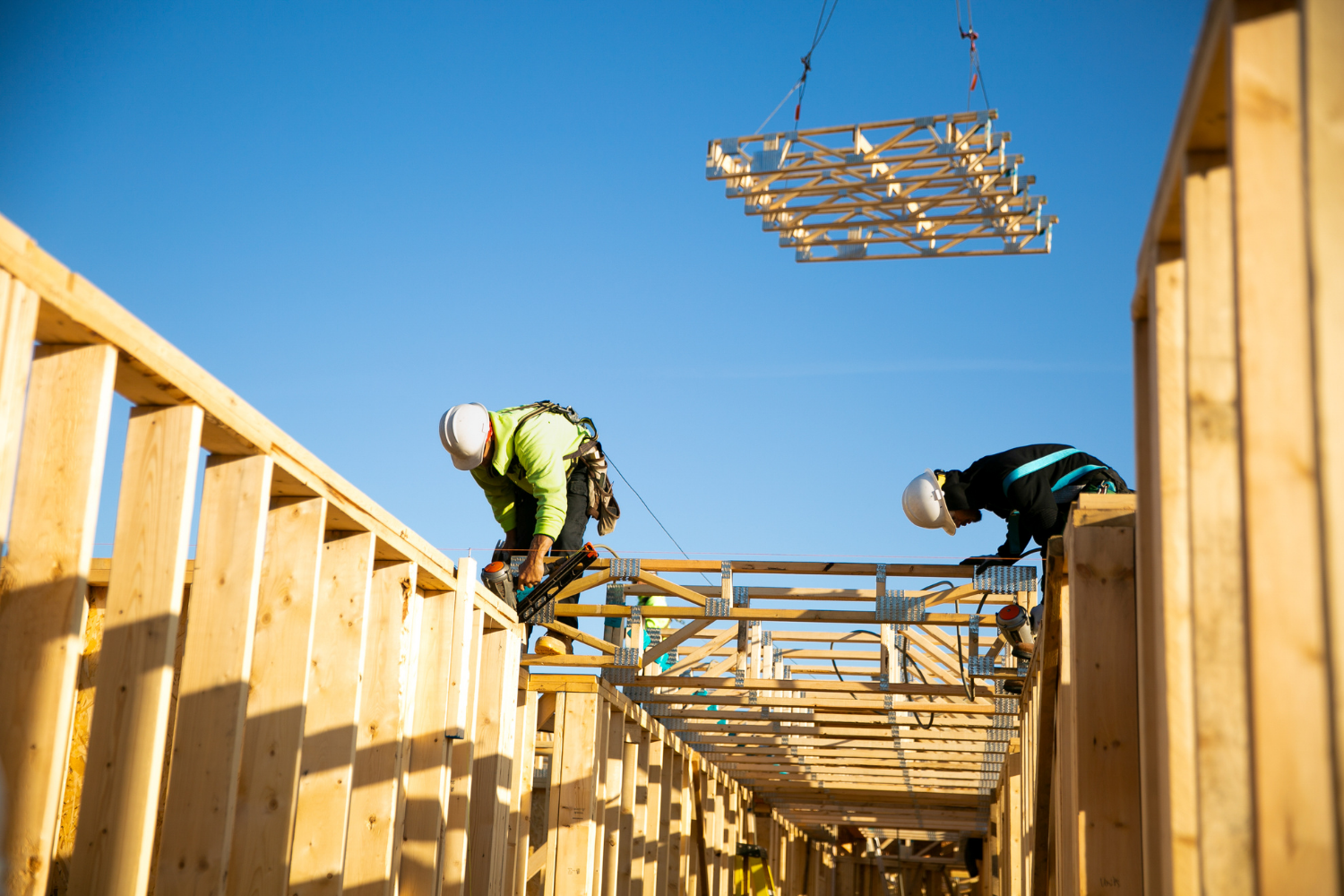 A QBC framer in full PPE nailing crane-set floor trusses into place on a multifamily project, with a second carpenter behind him and another truss being lifted in the background