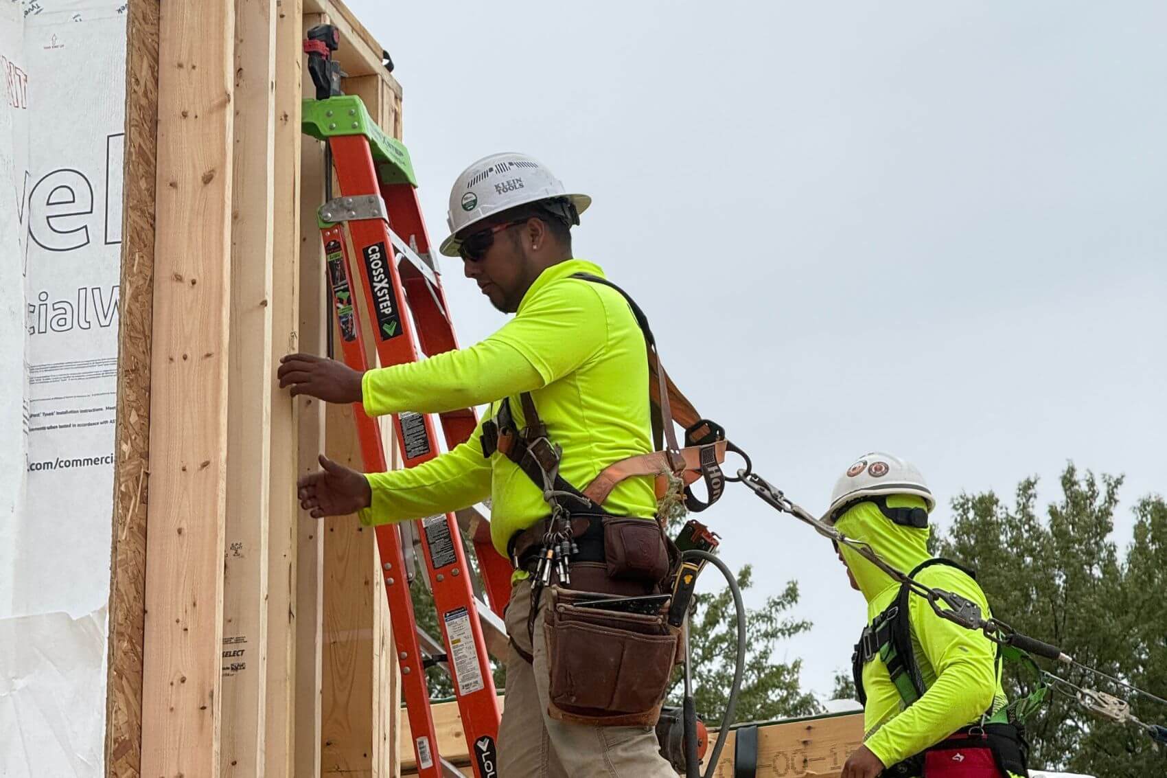 A framing carpenter in full safety harness gear and PPE sets a wall on a multifamily platform build in DeForest, Wisconsin.