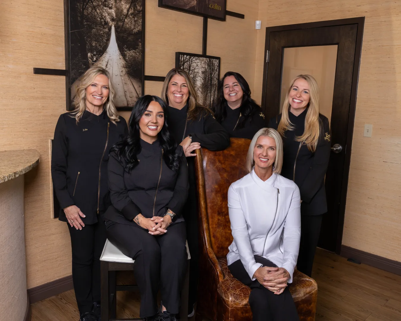 Six smiling women in black and white uniforms posing indoors with wood-paneled walls and framed nature photos. Simspon DDS Team