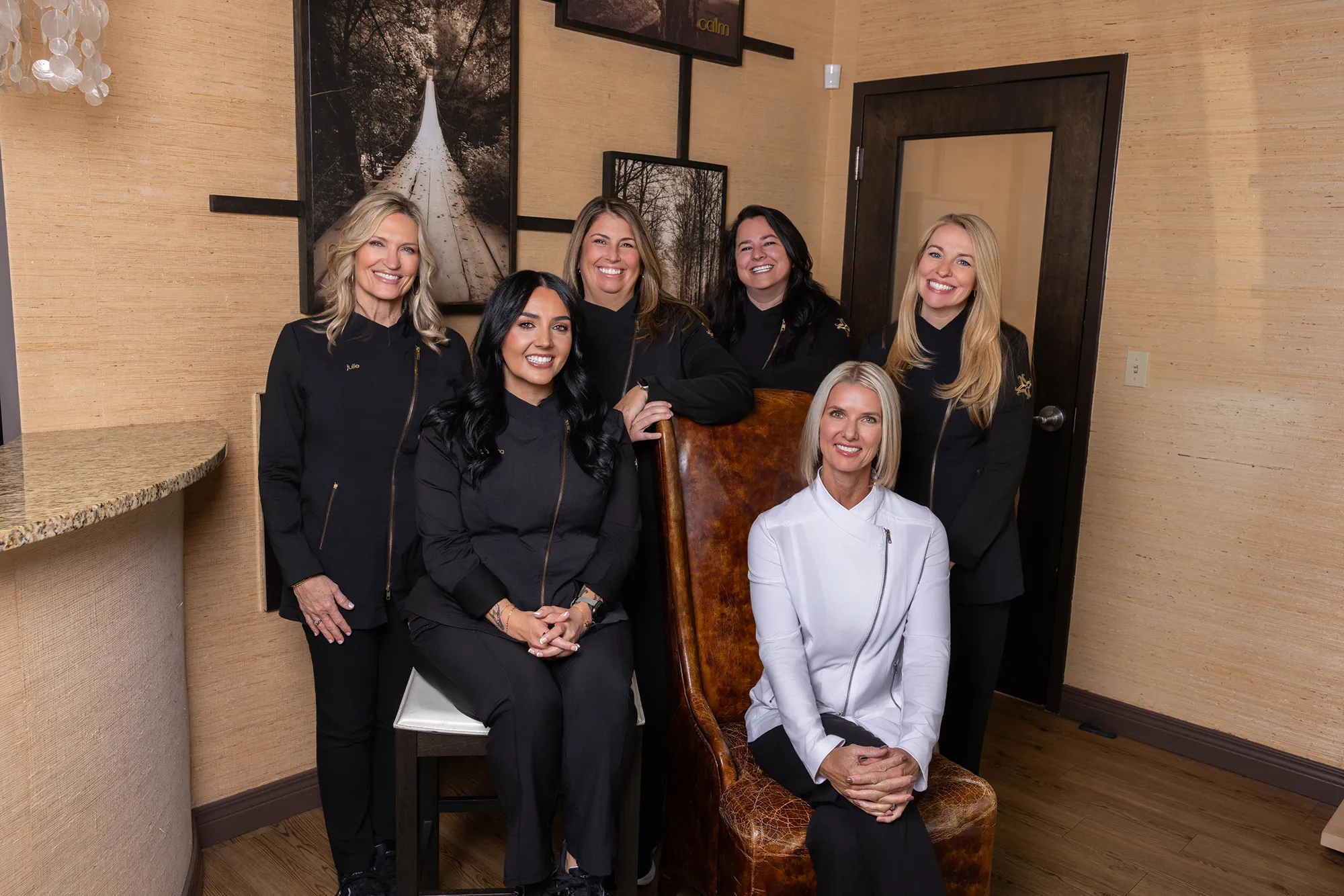 Group portrait of six smiling women in black and white professional uniforms posed in a warmly decorated office.
