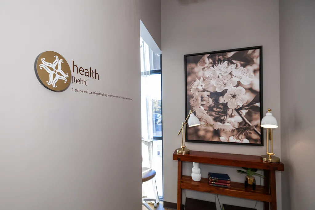 Interior wall with a health definition decal next to a wooden console table holding two brass and white lamps, a vase, books, a small plant, and a framed sepia floral photograph above.
