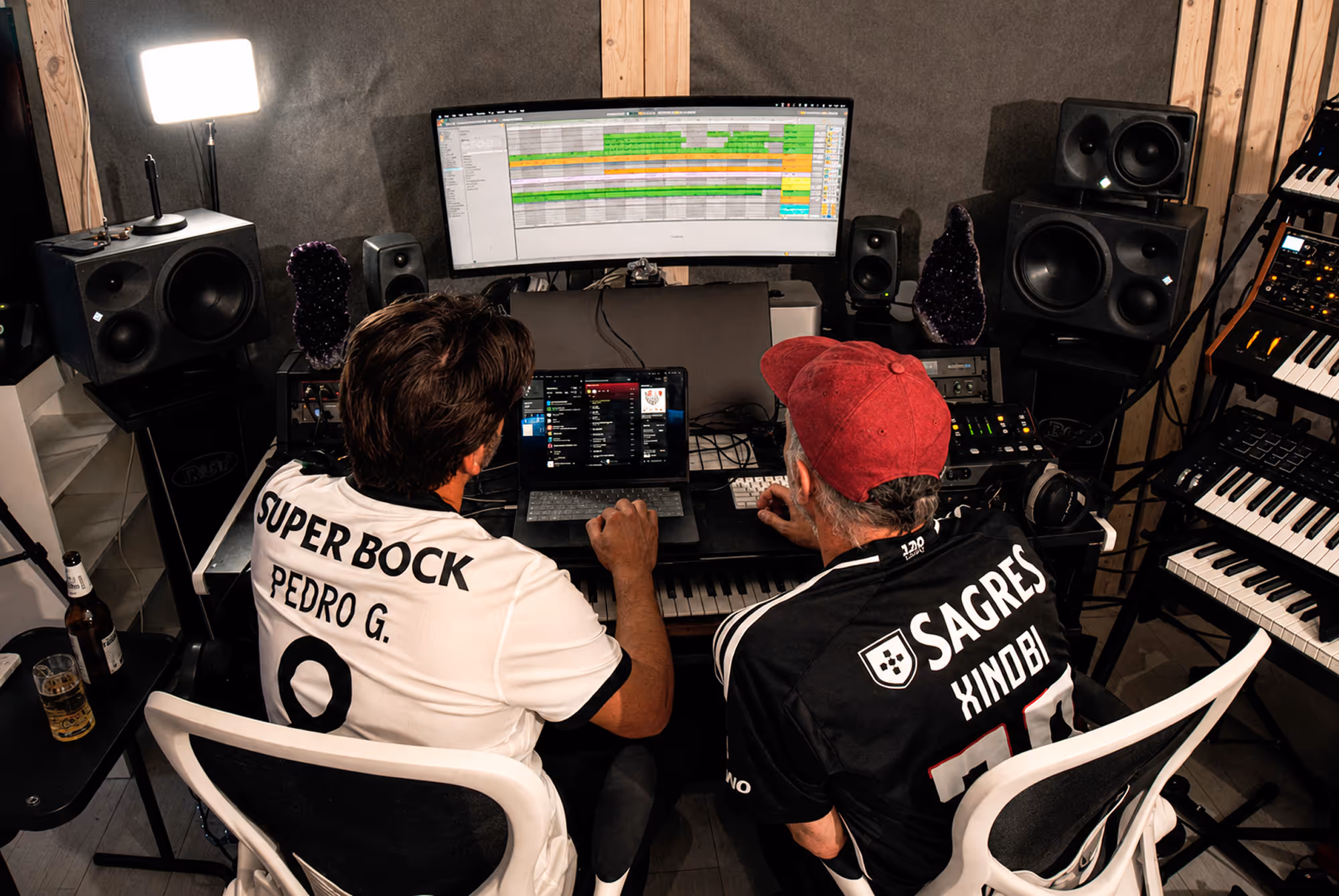 Two men in branded shirts working on music production with computers and keyboards in a studio.