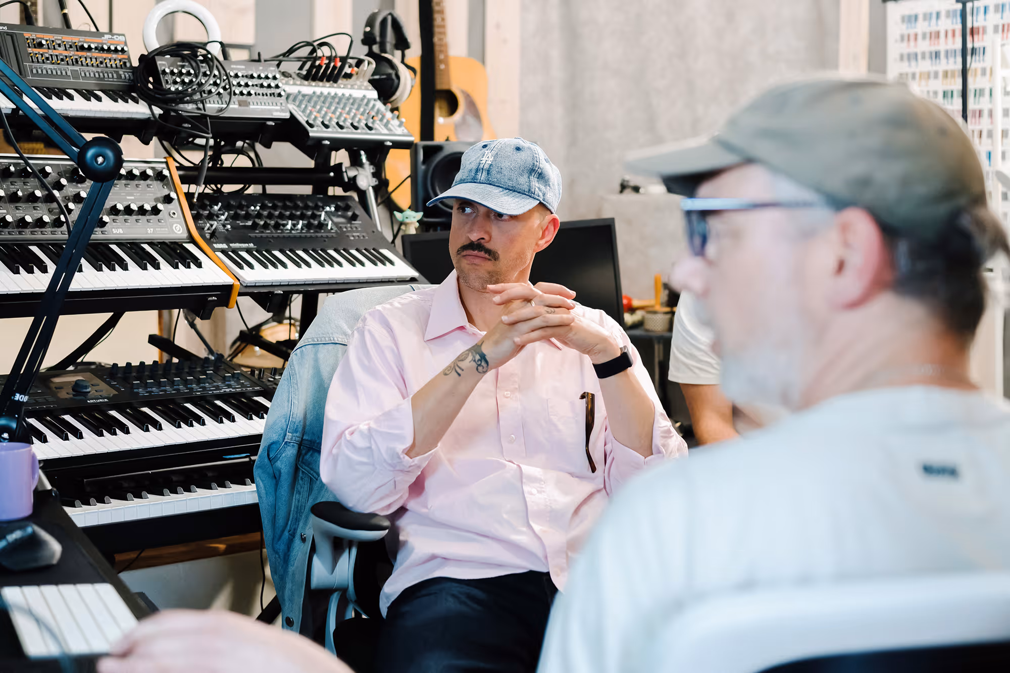 Man in a blue cap and pink shirt sitting in a music studio with keyboards and equipment behind him.