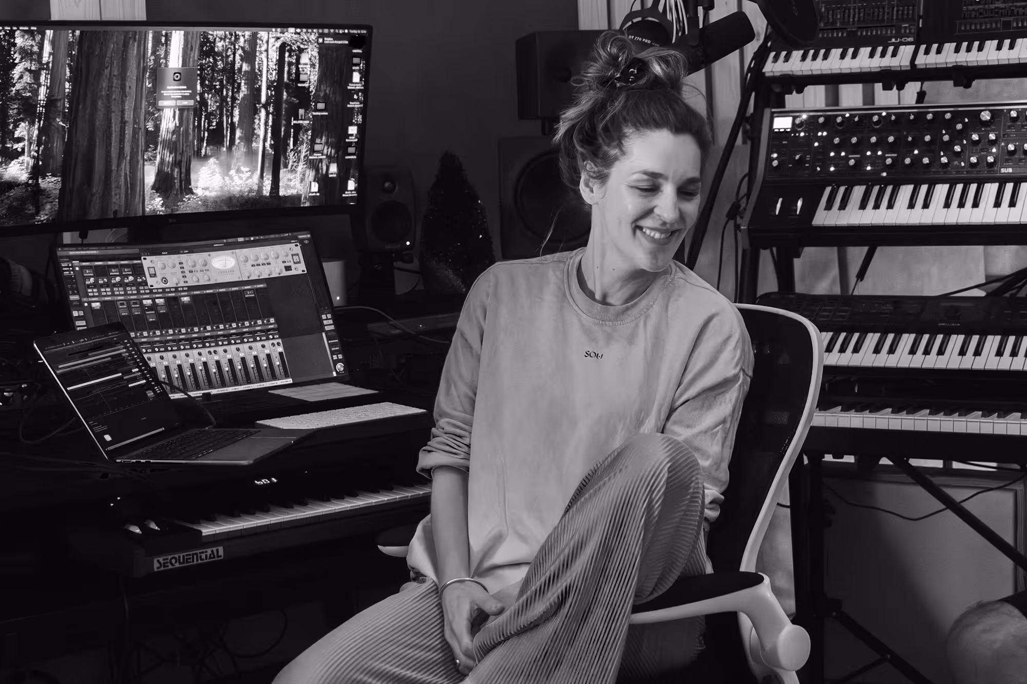 Smiling woman sitting in chair in a music studio surrounded by keyboards and computer screens.