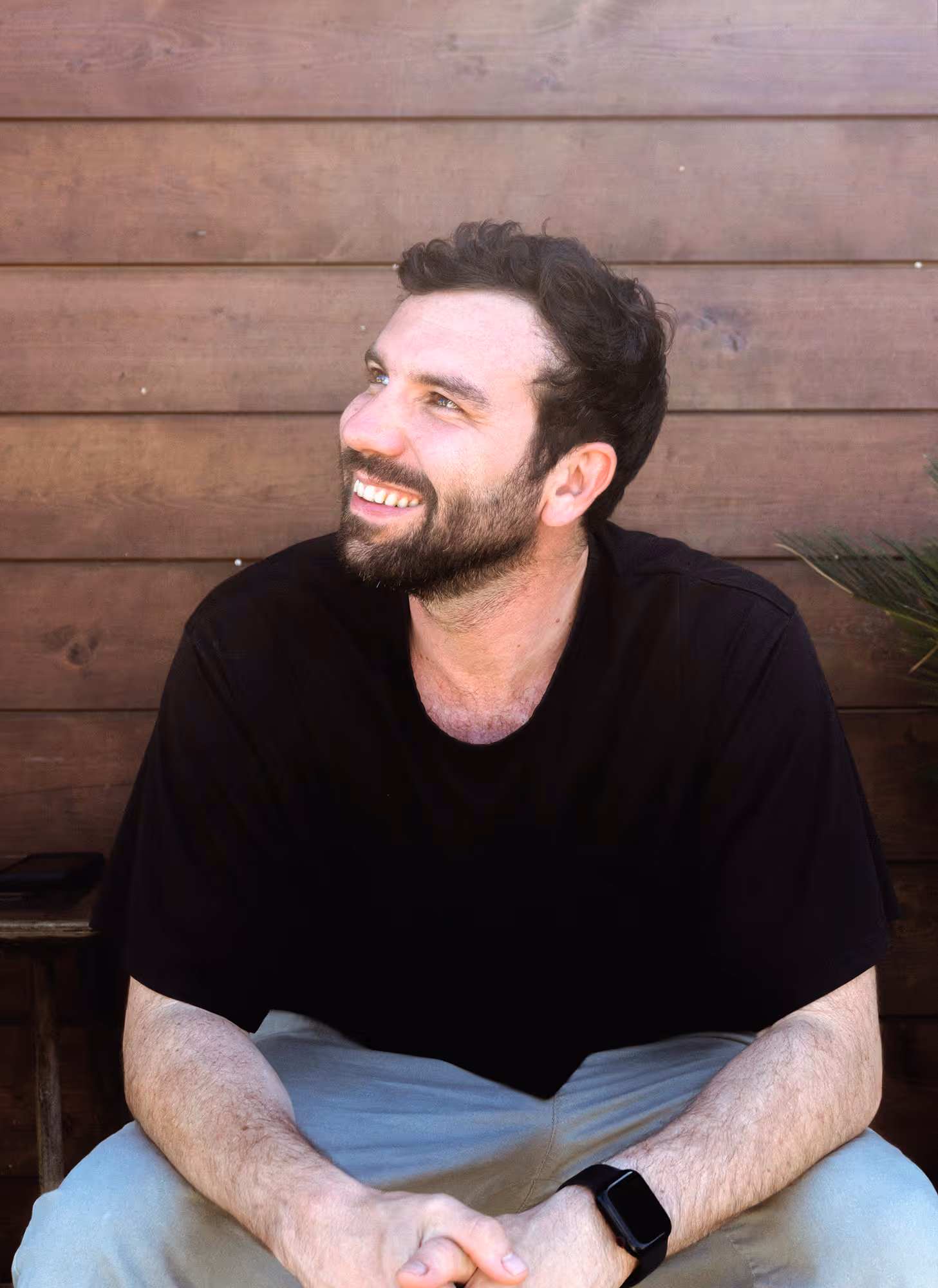 Bearded man in black shirt, light pants, and smartwatch smiling while sitting against wooden wall.