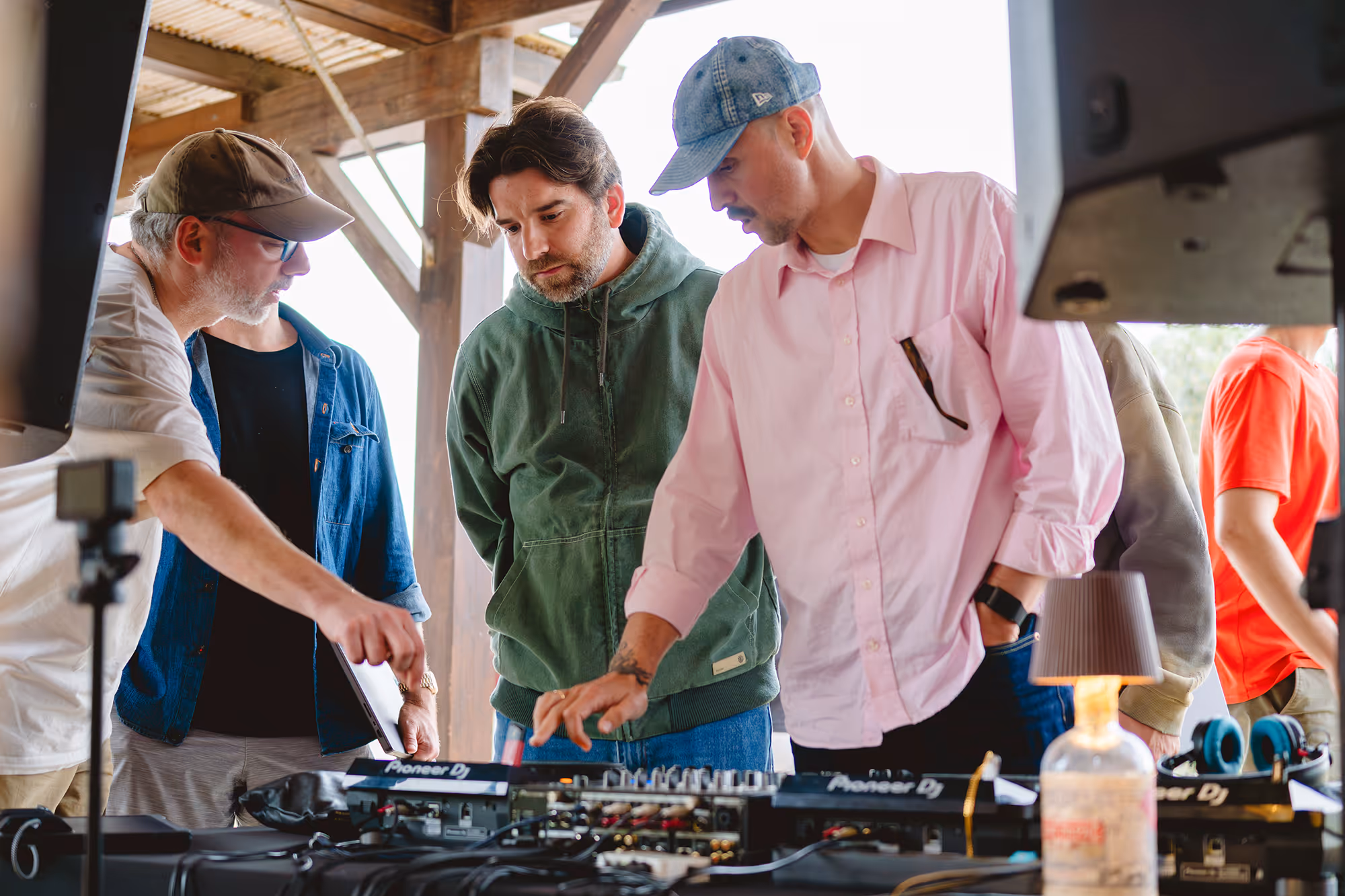 Three men focused on DJ equipment in a wooden outdoor structure, one adjusting controls.
