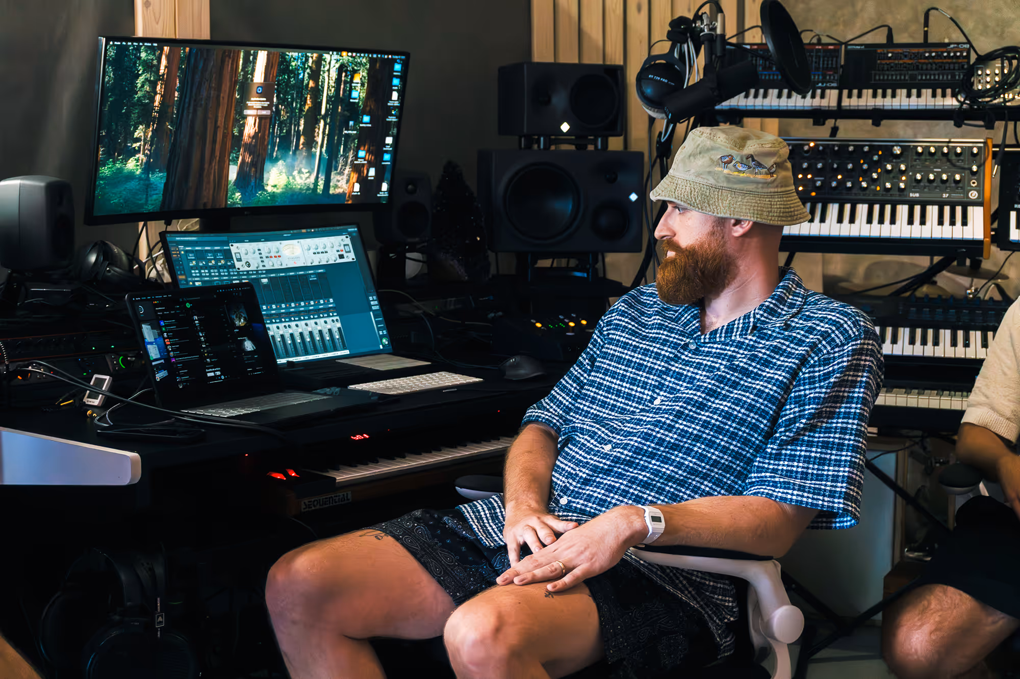 Bearded man in a bucket hat and plaid shirt sitting in a music studio with keyboards and monitors.