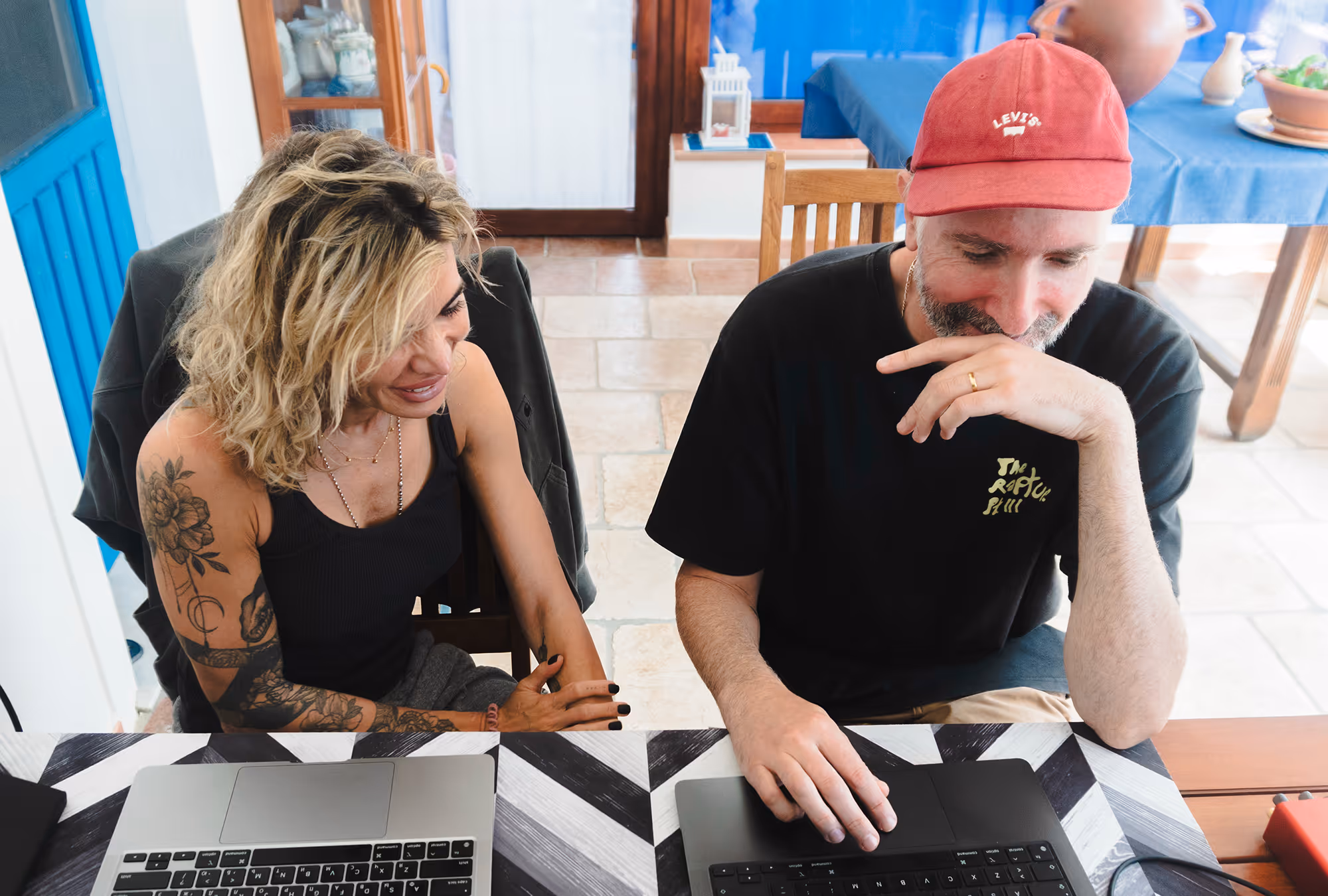 Man in red cap and woman with tattoos sit at a table working on laptops together indoors.