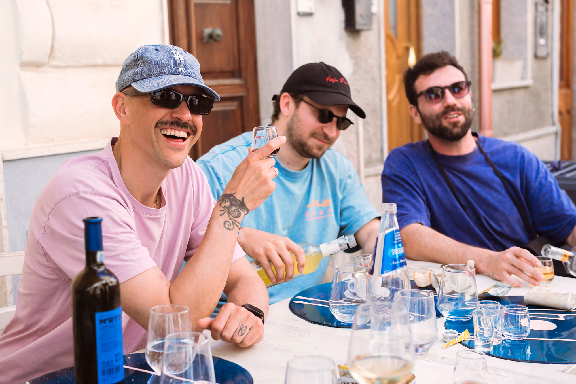 Three men wearing sunglasses sitting outdoors at a table with drinks, smiling and enjoying themselves.