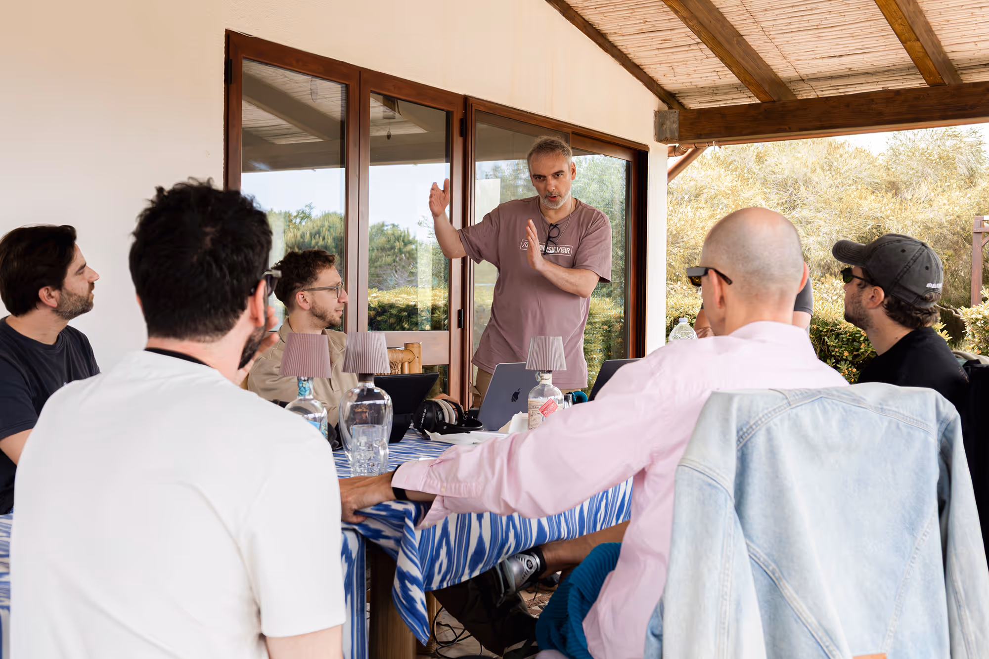 Man standing and gesturing while five seated men listen during an outdoor meeting.