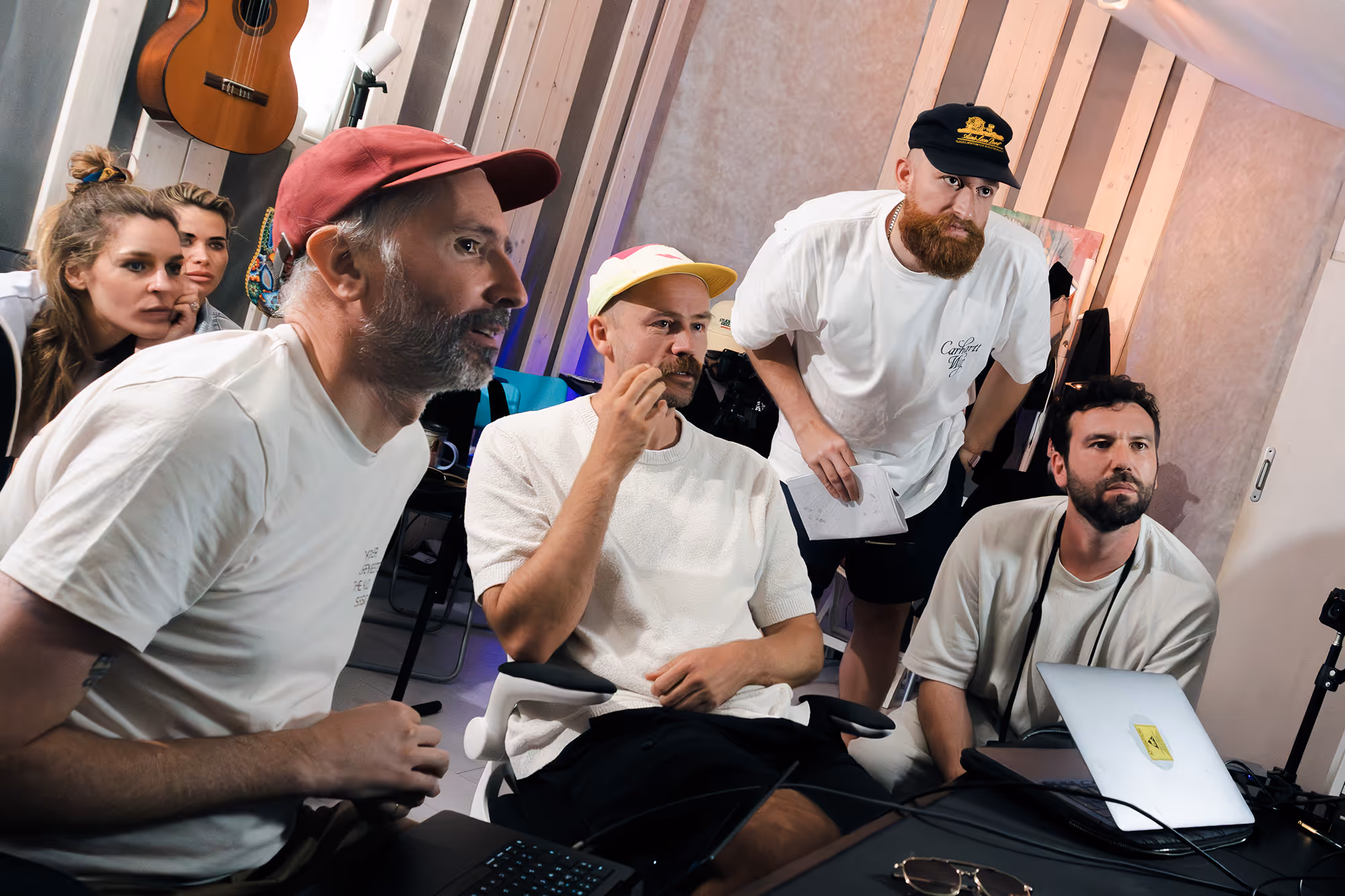 Five people focused on a laptop screen in a casual work setting with musical instruments in the background.
