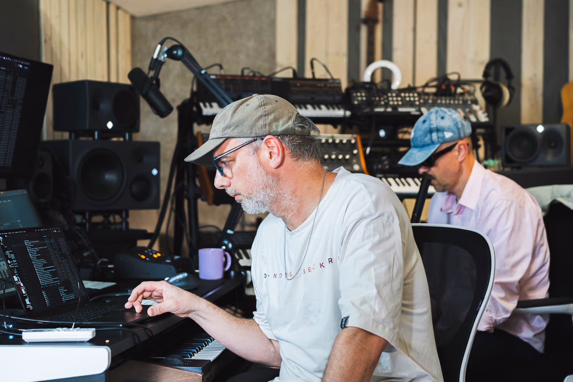 Two men working in a music studio with keyboards, speakers, and a laptop.