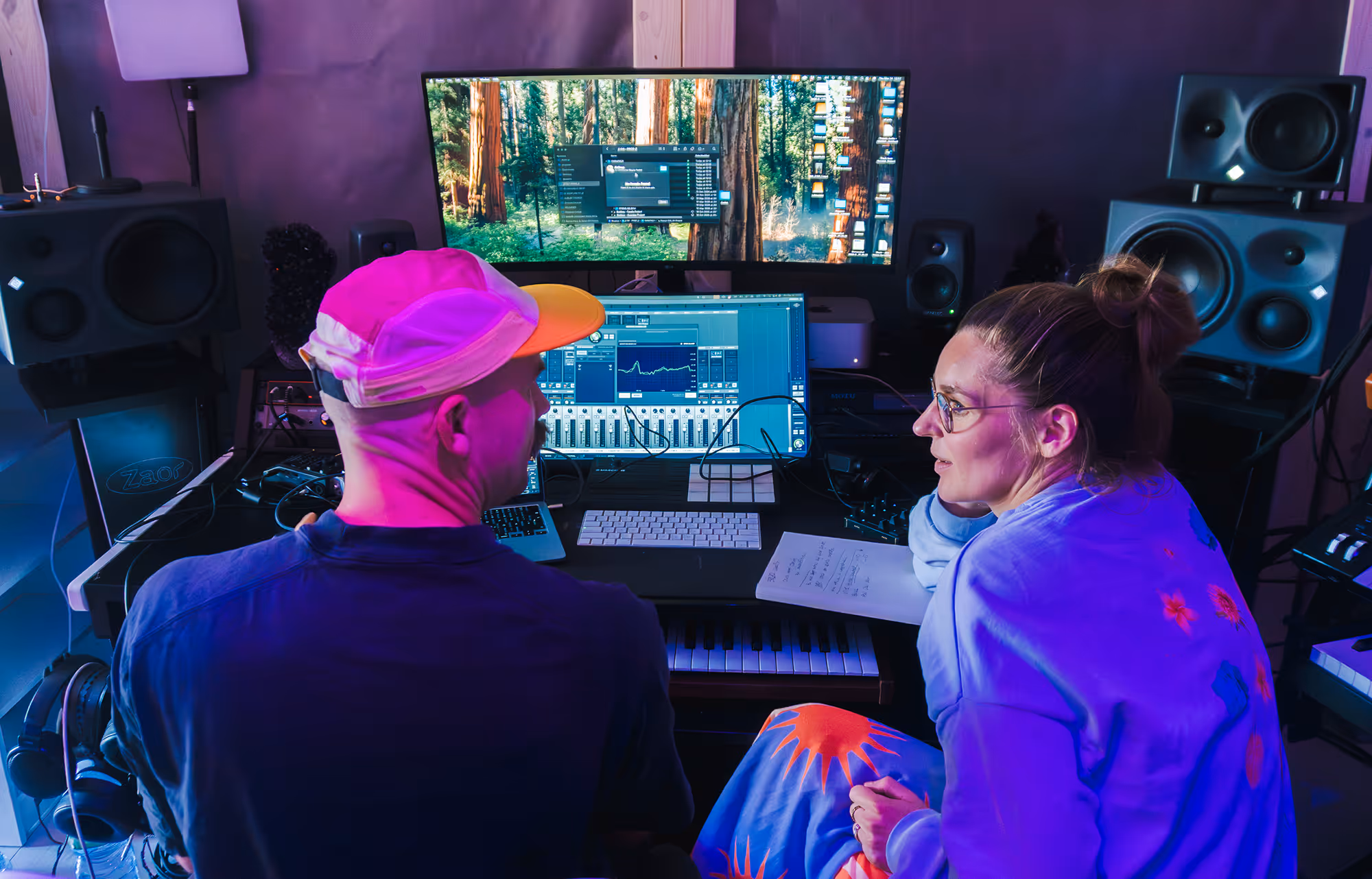 Two people in a recording studio with computer screens, speakers, keyboard, and audio equipment.