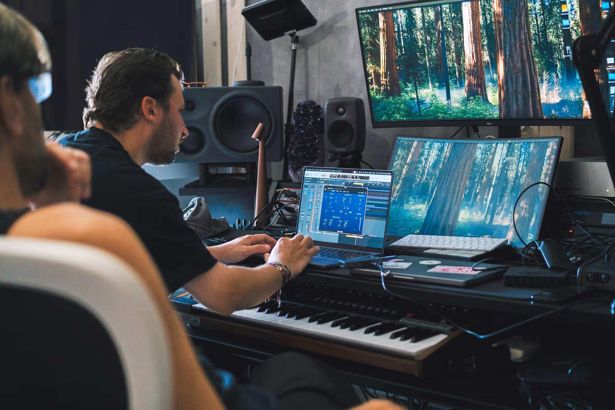 Man working on laptop with music software in a studio surrounded by monitors, keyboard, and speakers.