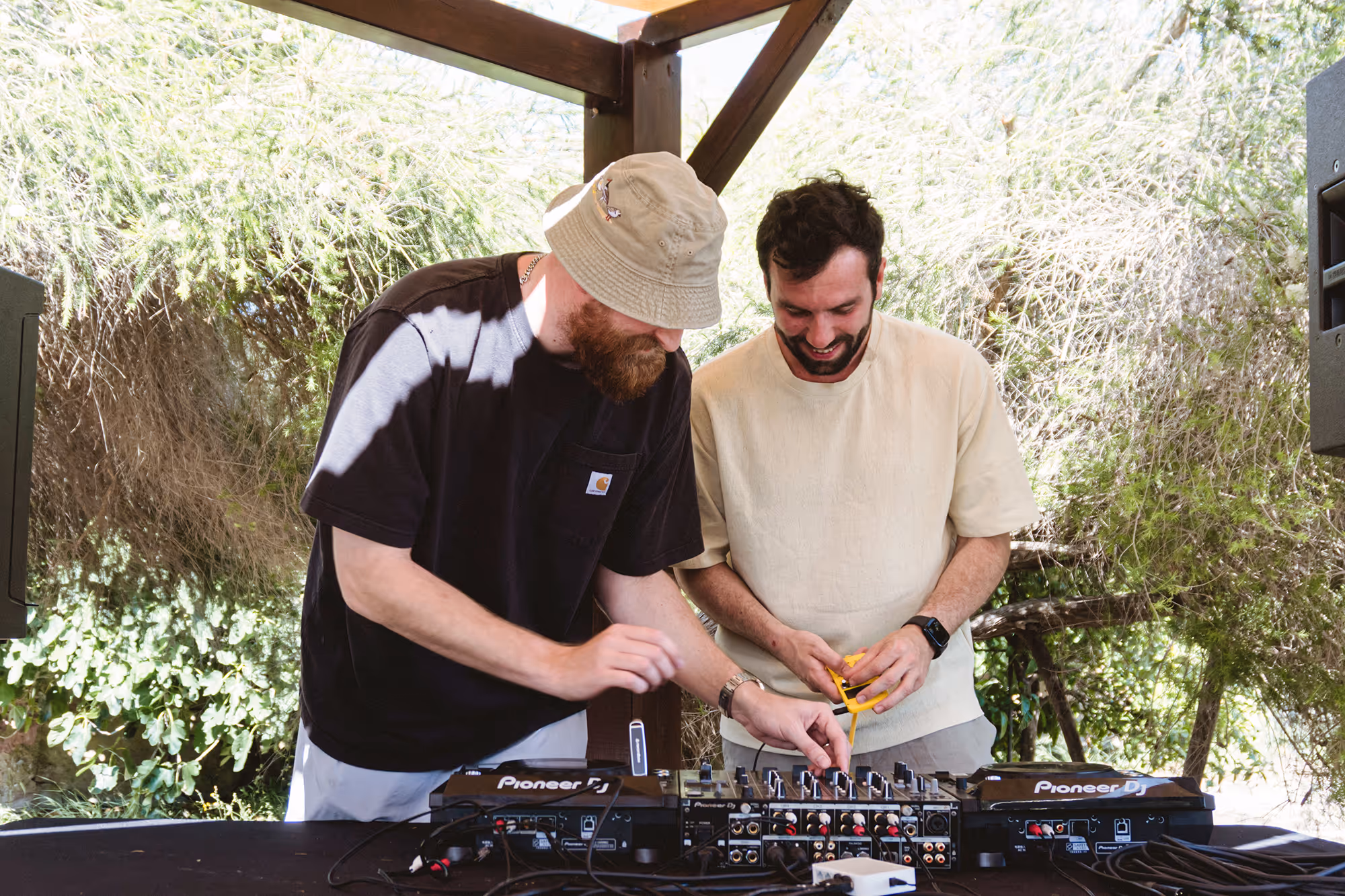 Two men operating DJ music mixers outdoors under a wooden shelter surrounded by greenery.