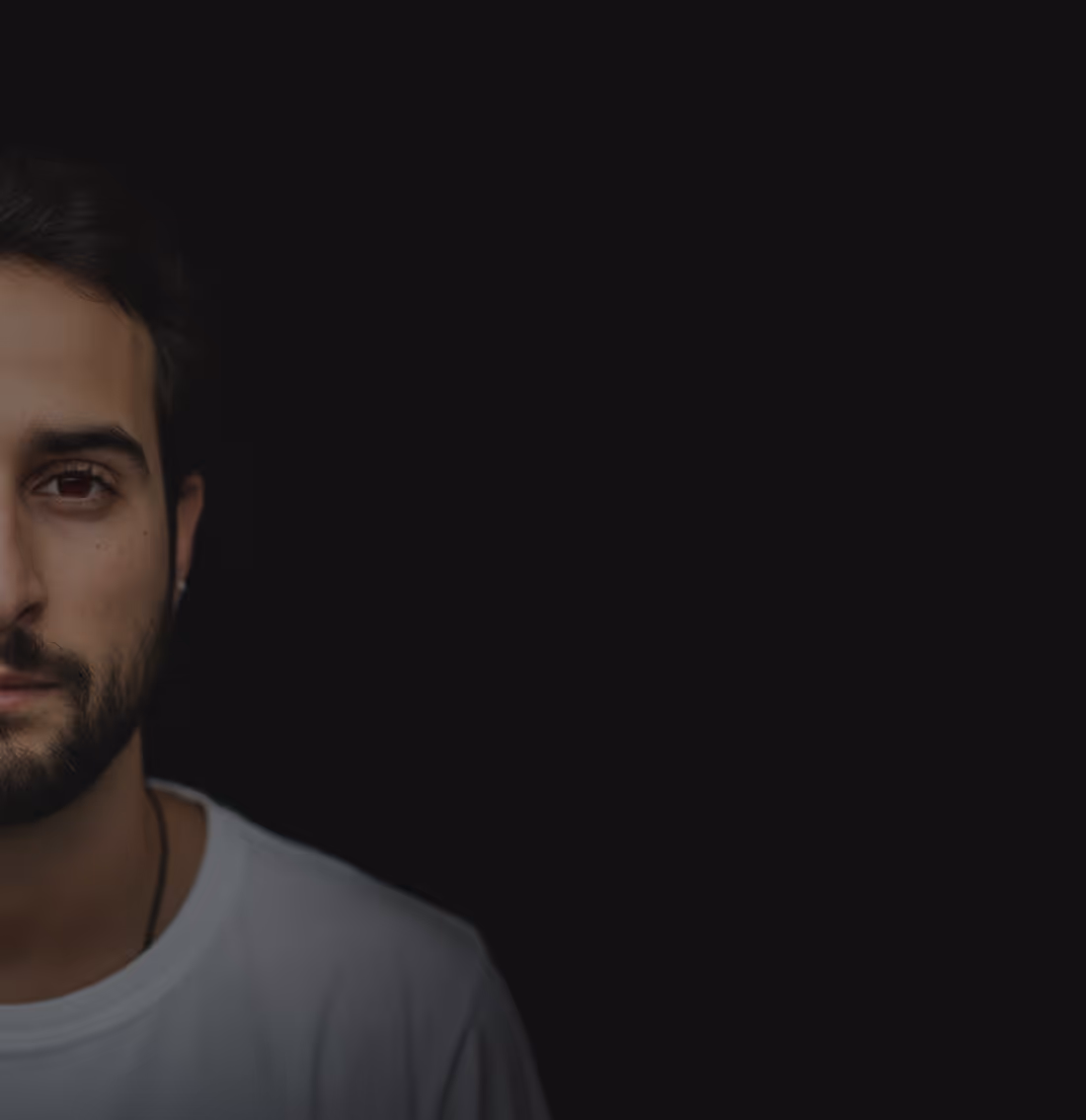 Close-up portrait of a young man with dark hair and beard wearing a white shirt against a black background, showing only the left half of his face.