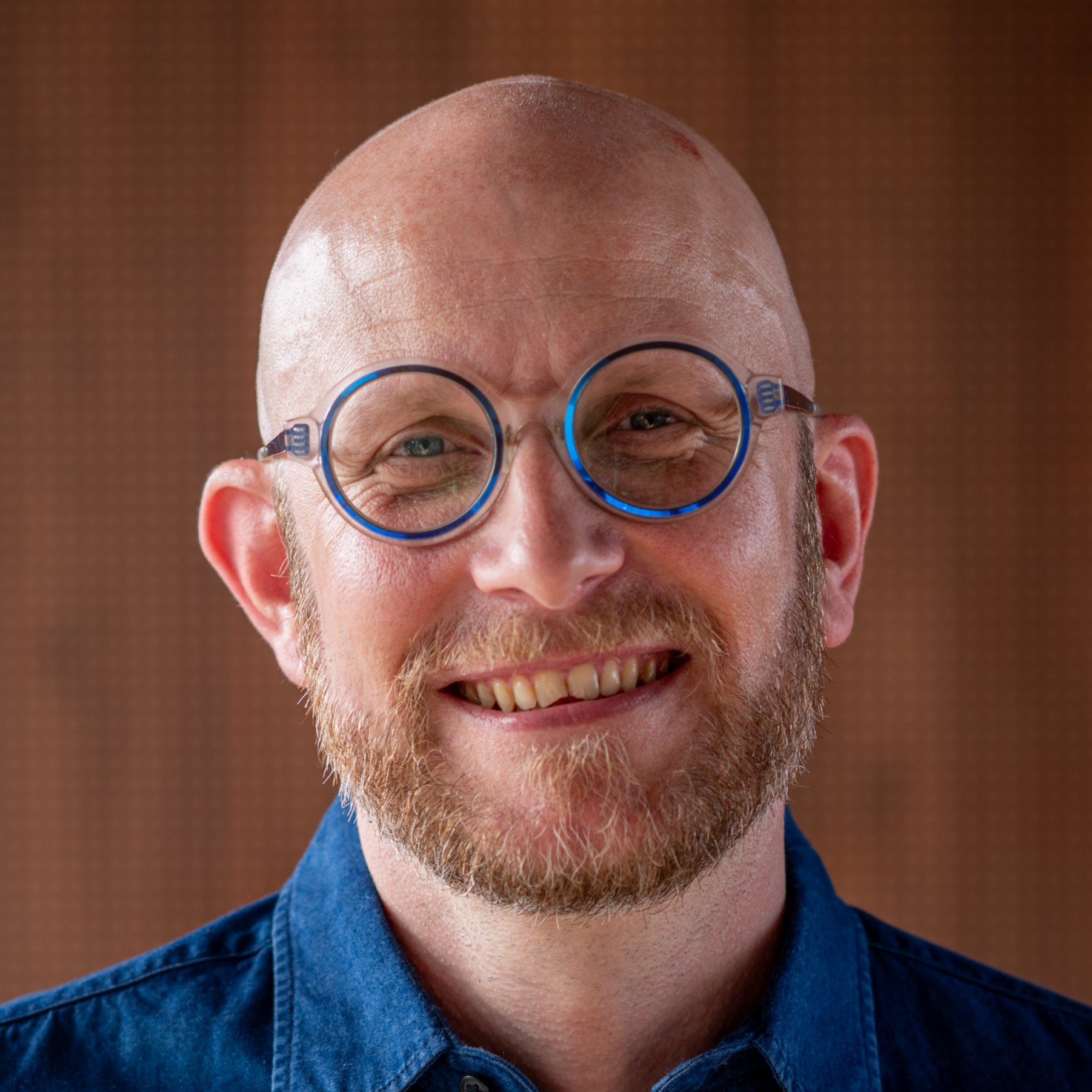 Homme chauve avec lunettes rondes à monture bleue, barbe rousse et chemise en denim souriant.
