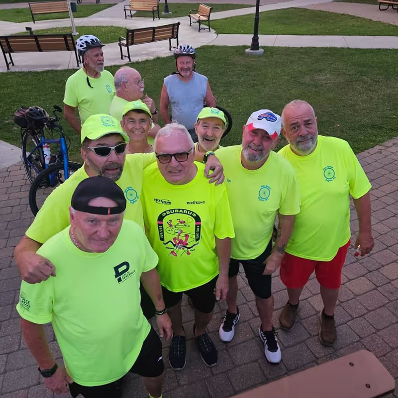 A cheerful group of cyclists in neon yellow shirts gather for a group photo, smiling together in a park setting. Some are wearing bike helmets, enjoying a sunny day.