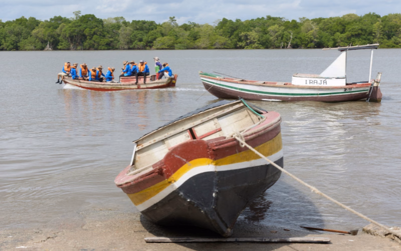 Two wooden boats on a calm river in Maranhão, Brazil, one carrying a group of people in orange life vests, with dense green mangrove forest along the far shore.