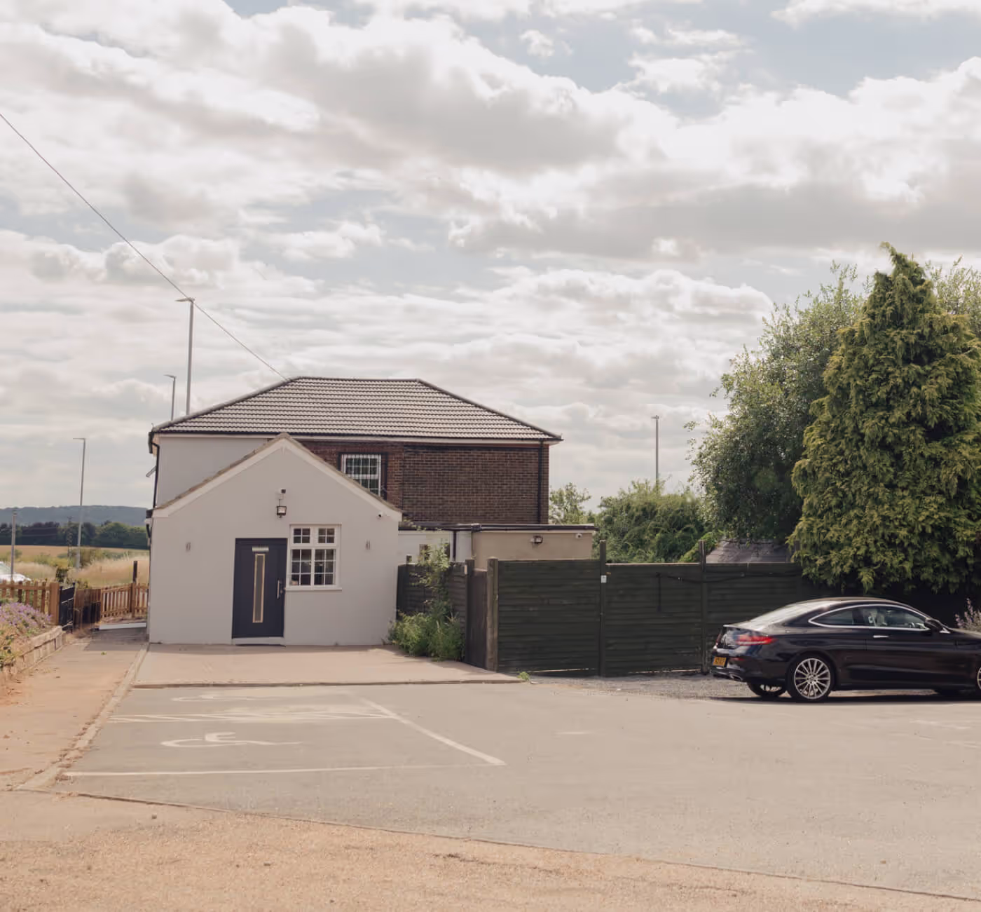Small white building with a dark door and window in front of a brick house, a black car parked beside, and cloudy sky above.