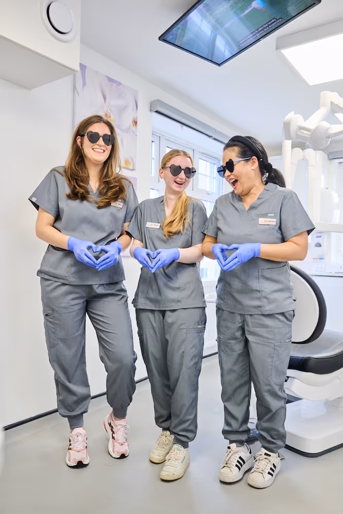 Three female dental professionals wearing gray scrubs and purple gloves, smiling and making heart shapes with their hands in a dental clinic.