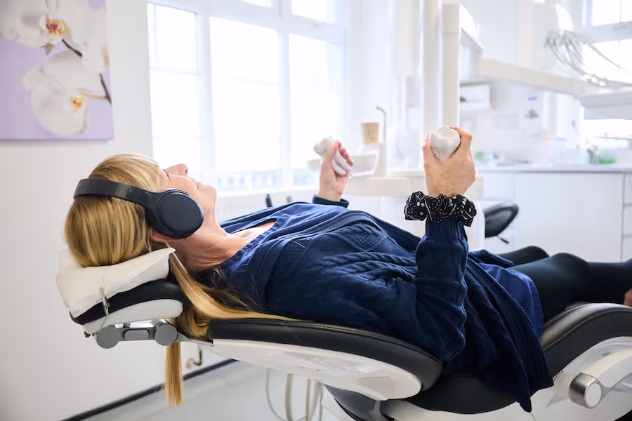 Woman lying back in a dental chair wearing headphones and squeezing two white stress-relief teeth toys.