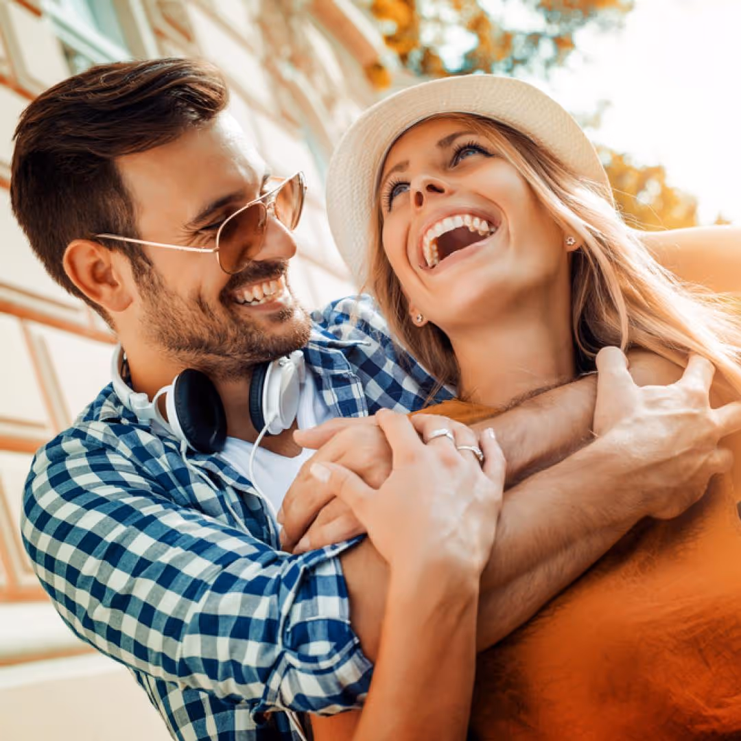 Smiling couple embracing happily outdoors, man wearing sunglasses and headphones, woman in white hat laughing.